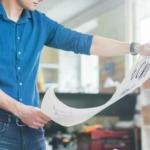 A person in a blue shirt examines a large sheet of printed signage with designs or drawings in a well-lit office or studio environment.