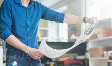 A person in a blue shirt examines a large sheet of printed signage with designs or drawings in a well-lit office or studio environment.
