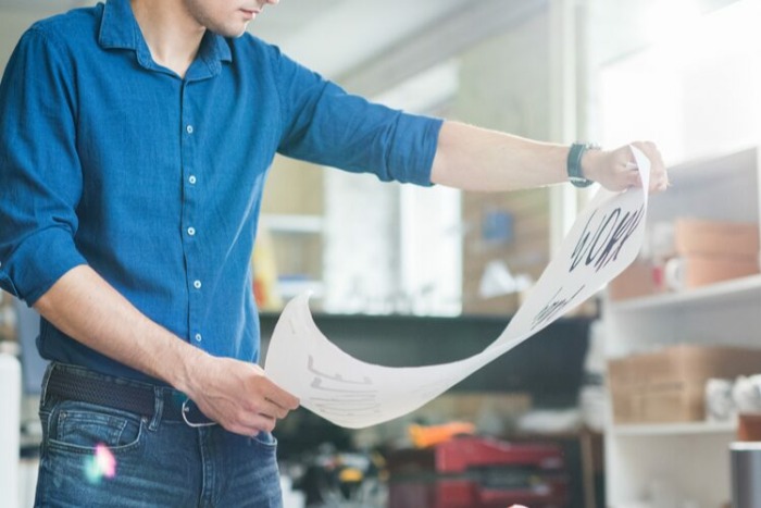 A person in a blue shirt examines a large sheet of printed signage with designs or drawings in a well-lit office or studio environment.